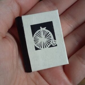 Miniature Bookbinding - 9th Dec 2023 A pale-skinned hand holding a miniature book with a black cloth spine and a cover with the image of a penny farthing on.